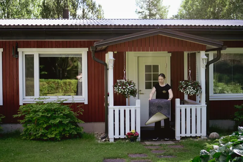 Pikkuapulainen puistelee mattoa punaisen omakotitalon kuistilla kesäpäivänä