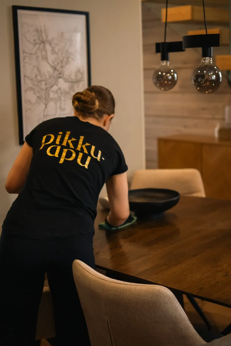 Pikkuapulainen pyyhkii ruokapöytää modernissa kodissa, työpaidan selässä kultainen pikku-apu-logo